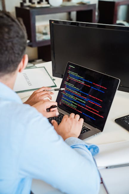 Photograph of a person working on a laptop with visible lines of colorful code on the screen, seated at a desk in a home or office environment. The desk has a black keyboard, a closed book labeled 'FU